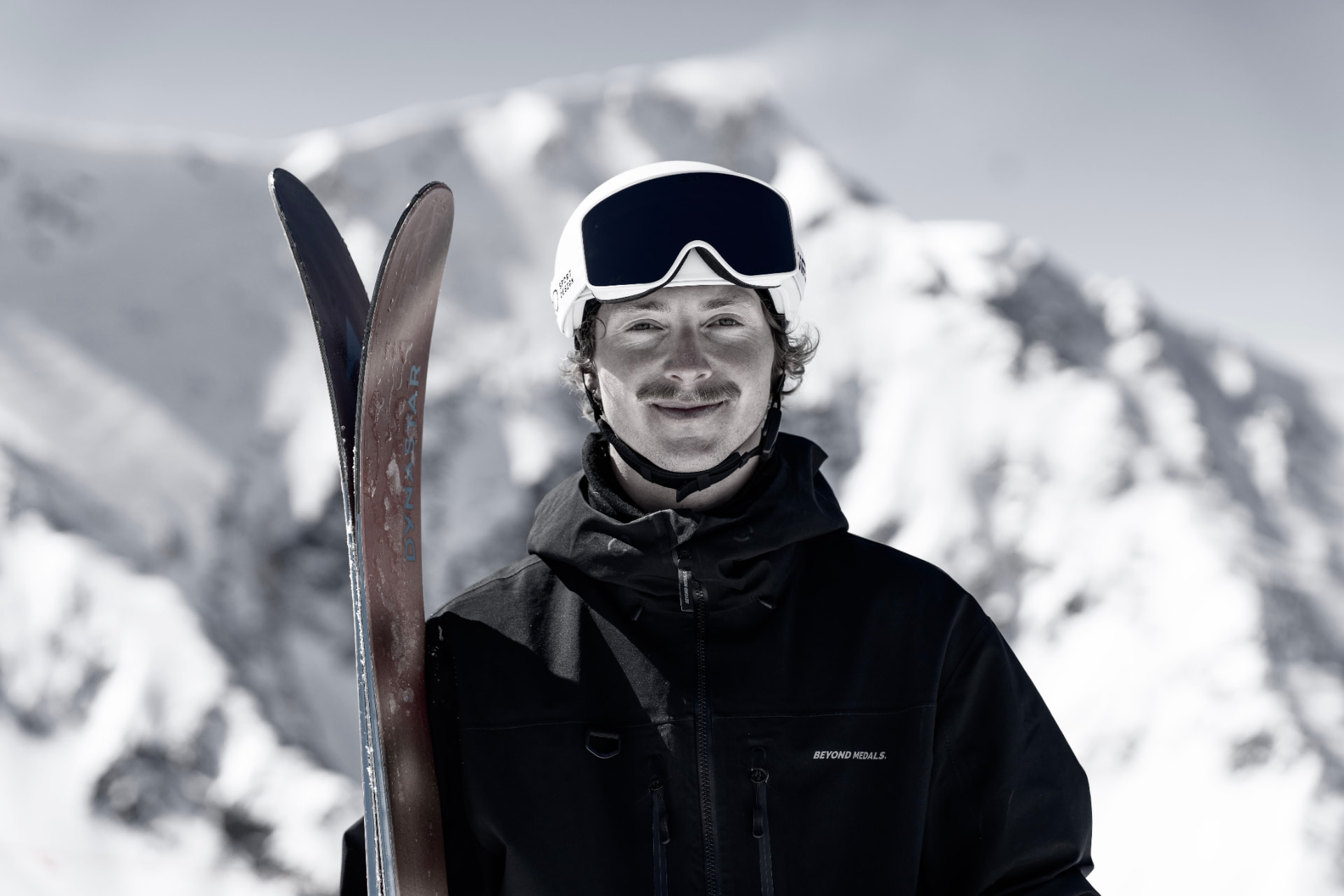 Portrait of Dillon Flinders in a helmet and goggles holding his skis, snow-covered peak behind him