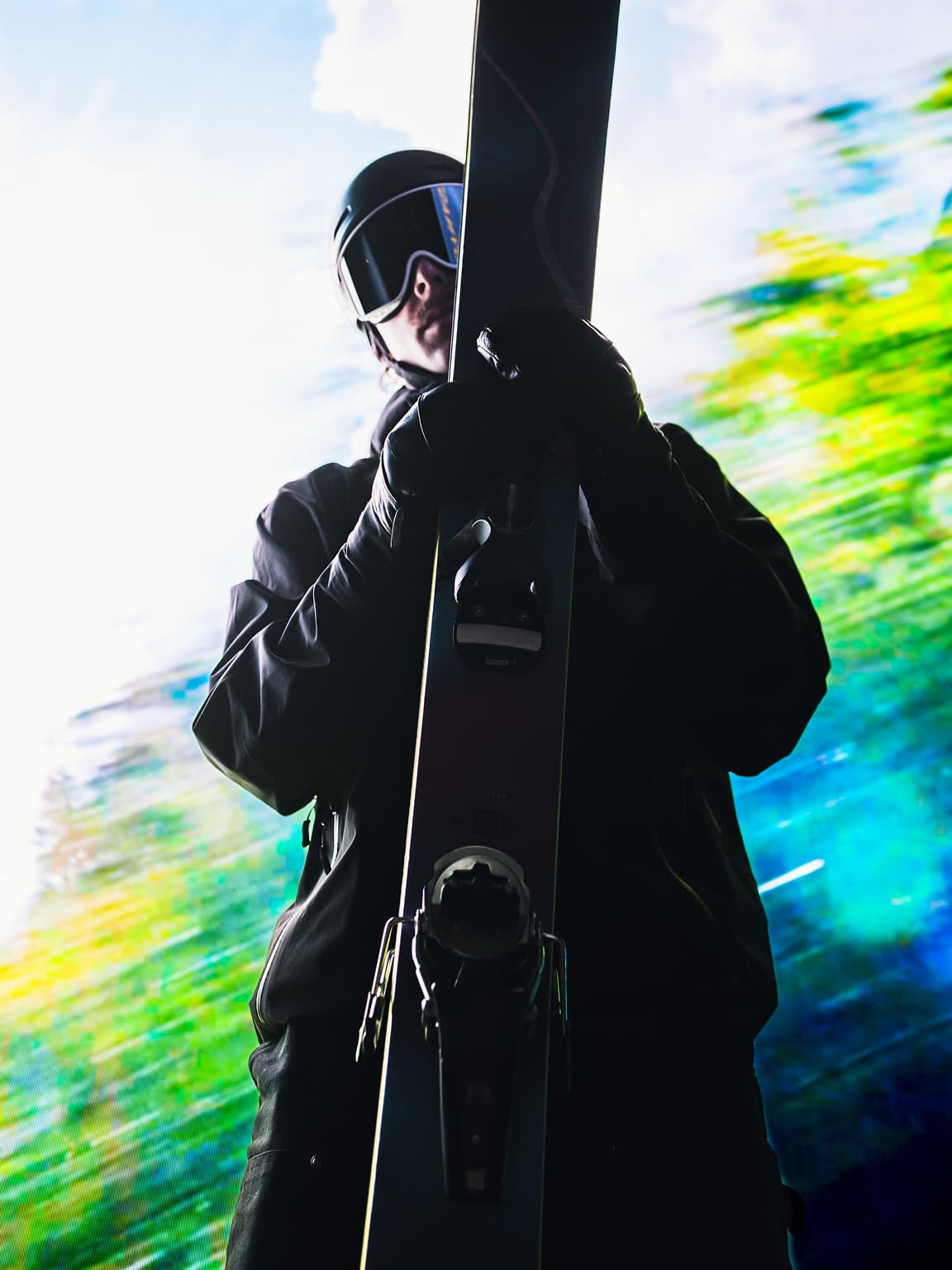 Dillon Flinders in ski gear holding a pair of skis vertically against a bright mountain backdrop